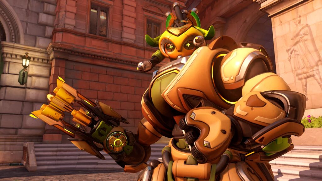 Orisa з Overwatch в золотих латах, націлившись зі зброї, стоячи на фоні міського дворика з червоною цеглою та кам'яними будівлями.